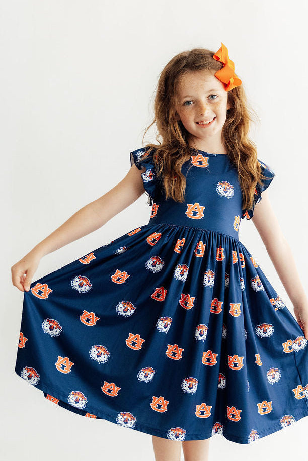 A young girl with long light brown hair wearing a navy blue dress with an orange and white tiger face pattern, holding out her skirt to show its volume. She is smiling and posing against a plain white studio background.