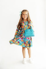 Young girl wearing a colorful floral dress holding a blue handbag on a white background