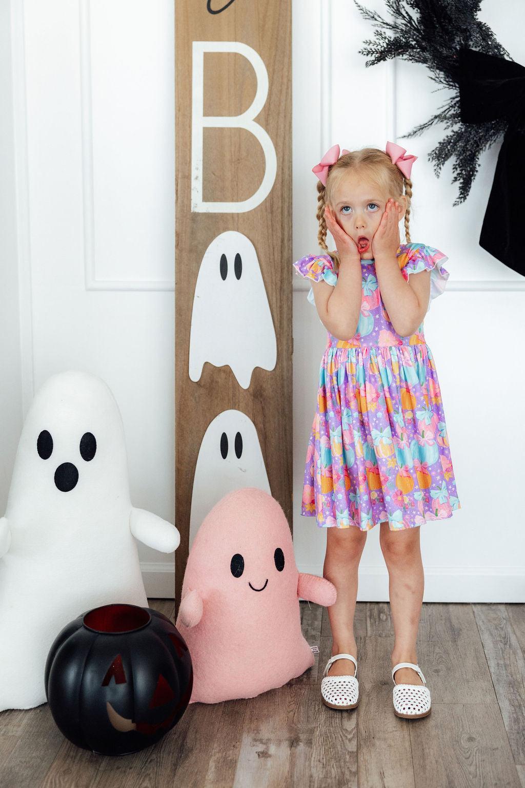 Child in a colorful dress standing next to Halloween-themed decorations including plush ghosts and a pumpkin.