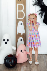 Child in a colorful dress standing next to Halloween-themed decorations including plush ghosts and a pumpkin.