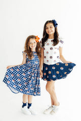 Two young girls modeling matching navy and white patterned outfits against a plain white studio background. The girl on the left wears a navy dress with white polka dots and small cartoon motifs, and the girl on the right wears a white top with navy cartoon-print shorts.