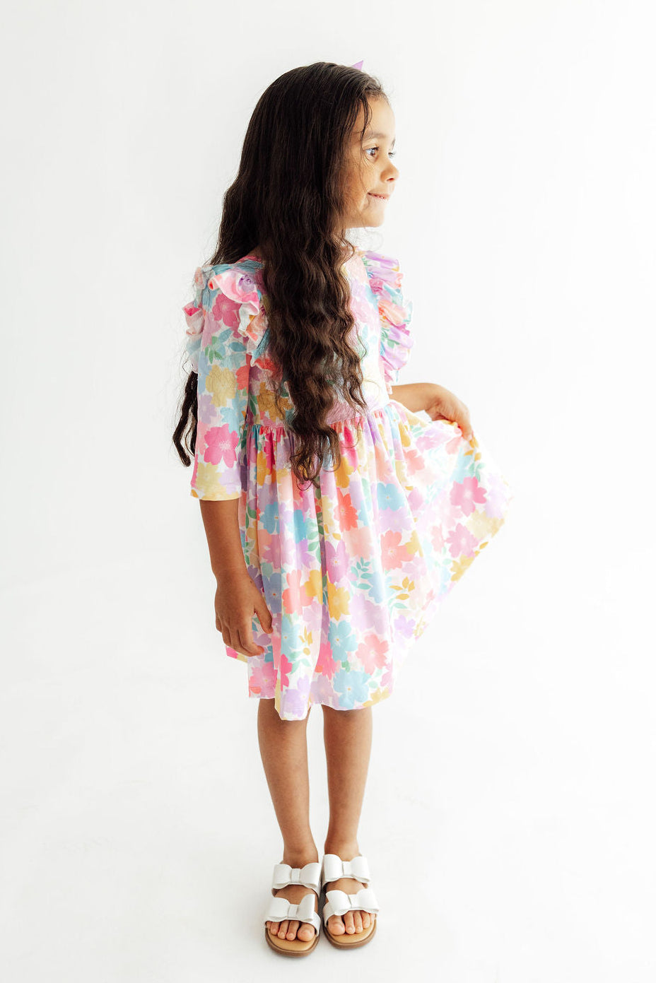 A young girl with long dark hair stands in profile wearing a pastel multicolor floral tiered dress with ruffled sleeves and white sandals, photographed against a plain white studio background.