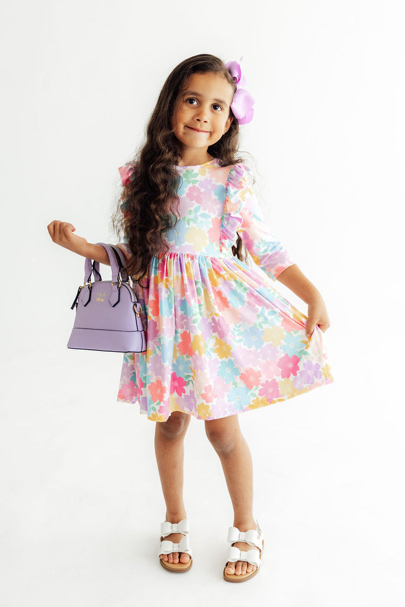 A young girl with long dark hair wearing a pastel multicolor floral dress, white sandals, and a purple flower hair clip, holding a lavender handbag against a plain white background.