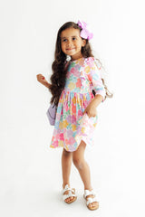 A young girl with long dark hair wearing a pastel multicolor floral print dress, a pink bow, and white sandals, standing against a plain white studio background.