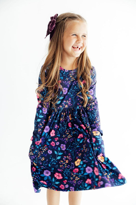 A young girl with long light brown hair wearing a dark purple long-sleeve dress with a colorful floral pattern, smiling and looking to the side against a plain white background.