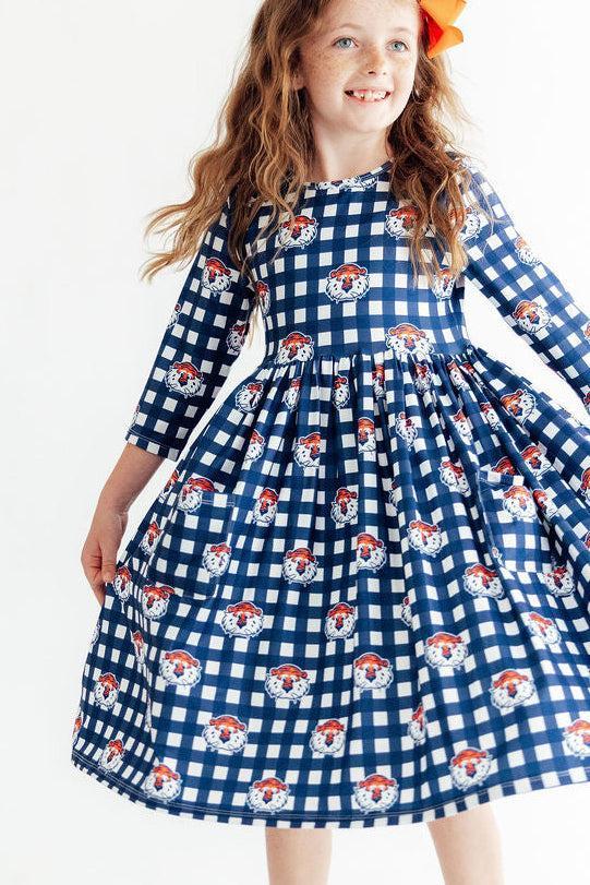 A young girl with long wavy light brown hair wearing a blue and white gingham dress with a repeating pattern of cartoon dog faces, posed against a plain white background.