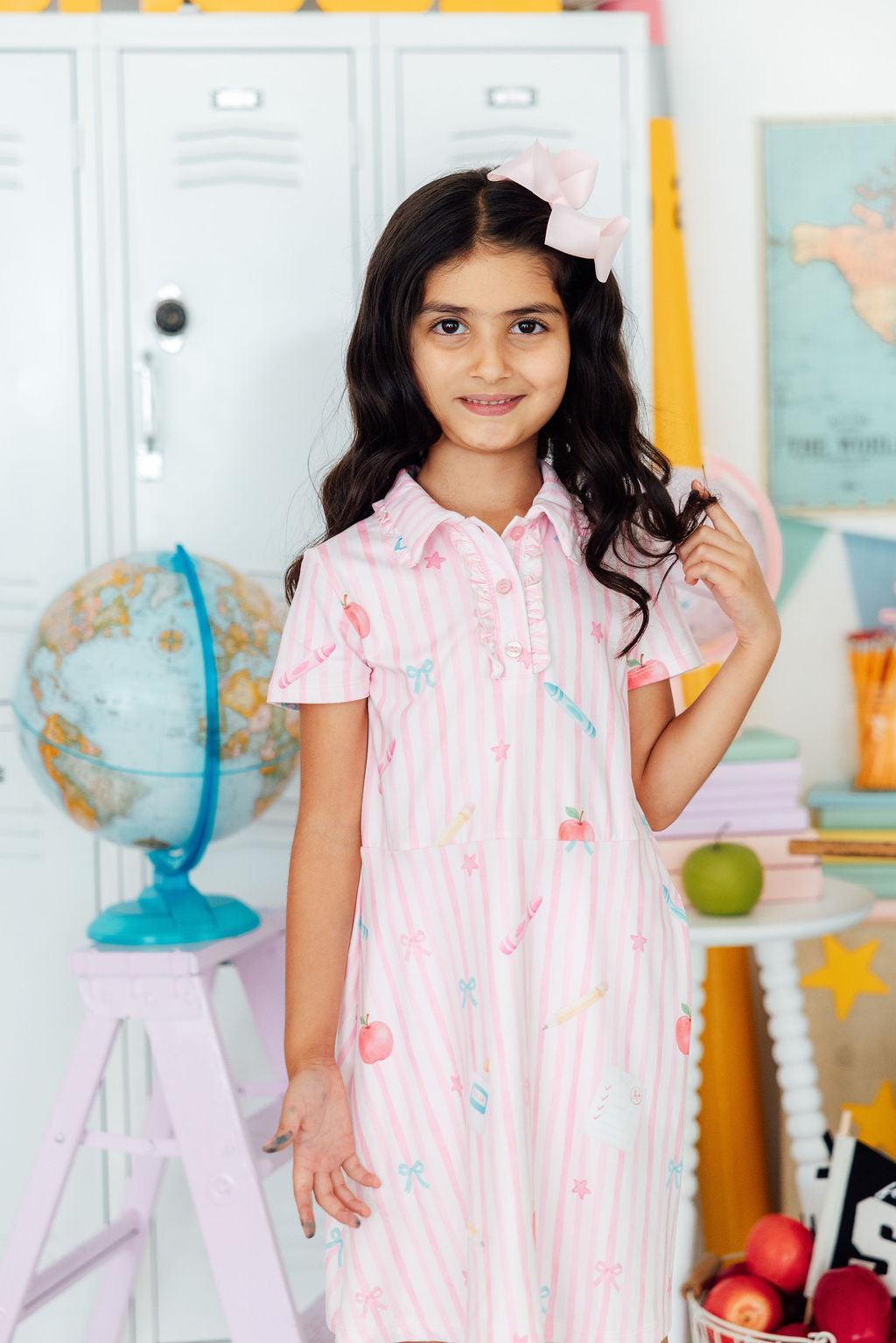 Young girl in a pink dress standing in a classroom setting with educational decor.