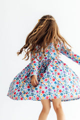 A young girl with long brown hair is shown from behind, wearing a long-sleeved, smocked-bust dress with a colorful floral print on a light blue background, captured mid-spin against a plain white studio background.