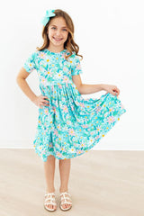 A young girl with a turquoise hair bow is wearing a short-sleeve, knee-length dress with a colorful floral and leaf print on a turquoise background. She is standing in a room with a white wall and light wood floor, holding out one side of the dress to show its flow.