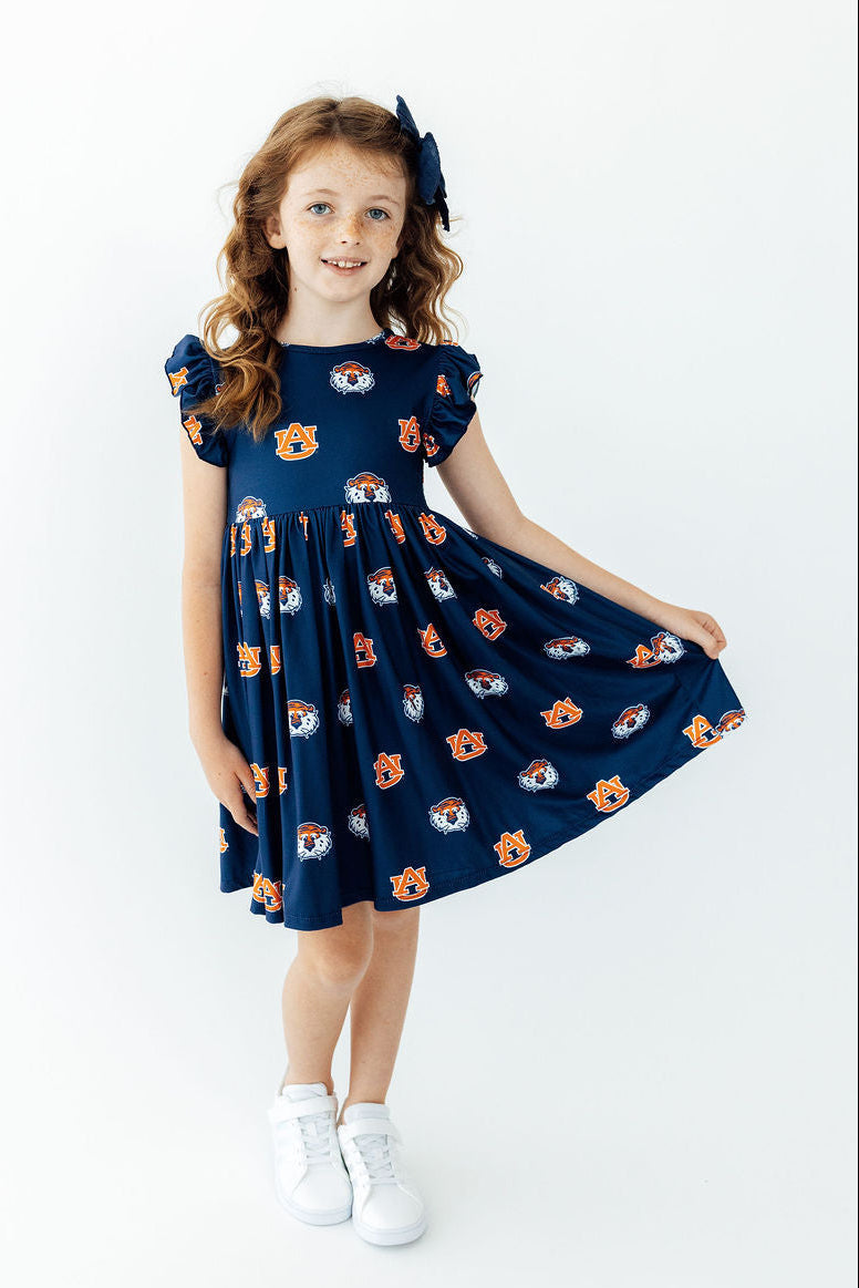 A young girl with wavy light brown hair wearing a navy blue dress with an orange and white cartoon bear pattern, white sneakers, and a matching navy hair bow, standing against a plain white background.