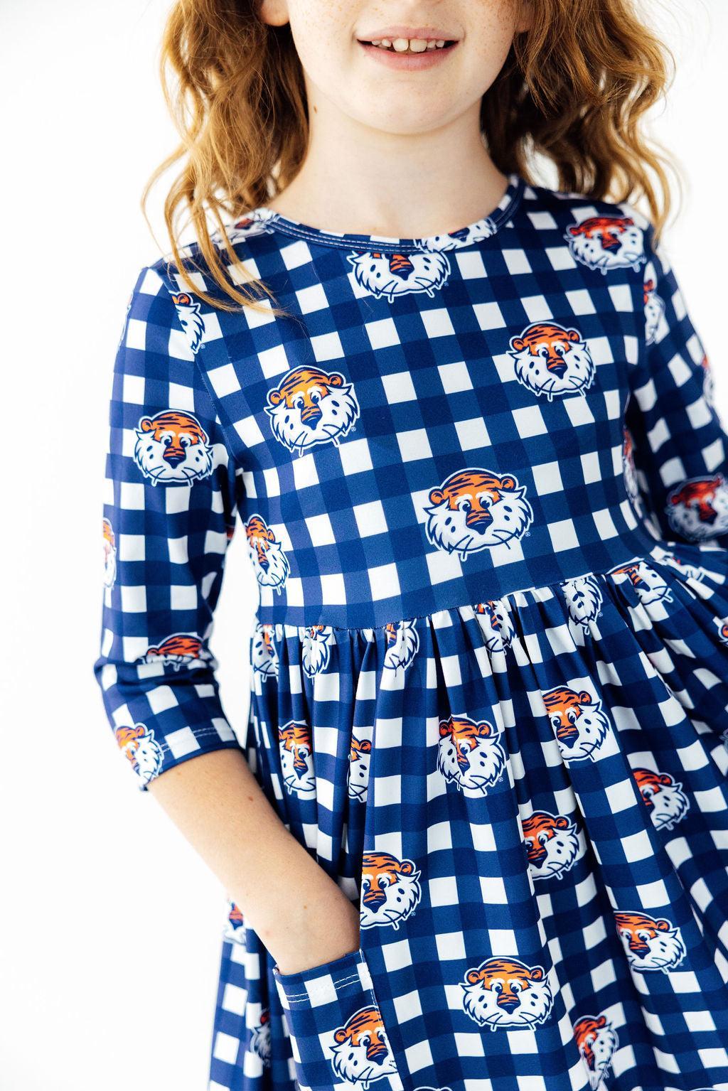 A young girl wearing a blue and white gingham dress with orange and white tiger mascot prints, shown from the shoulders down against a plain white background.