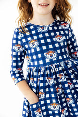 A young girl wearing a blue and white gingham dress with orange and white tiger mascot prints, shown from the shoulders down against a plain white background.