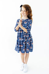 A young girl with curly light brown hair wearing a blue and white gingham dress with cartoon dog prints, white sneakers, and a black bow, standing against a solid white background.