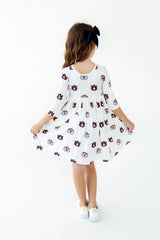 A young girl viewed from behind wearing a white three-quarter sleeve dress with a repeating cartoon cat face pattern, holding out her skirt to show its shape, against a plain white background.