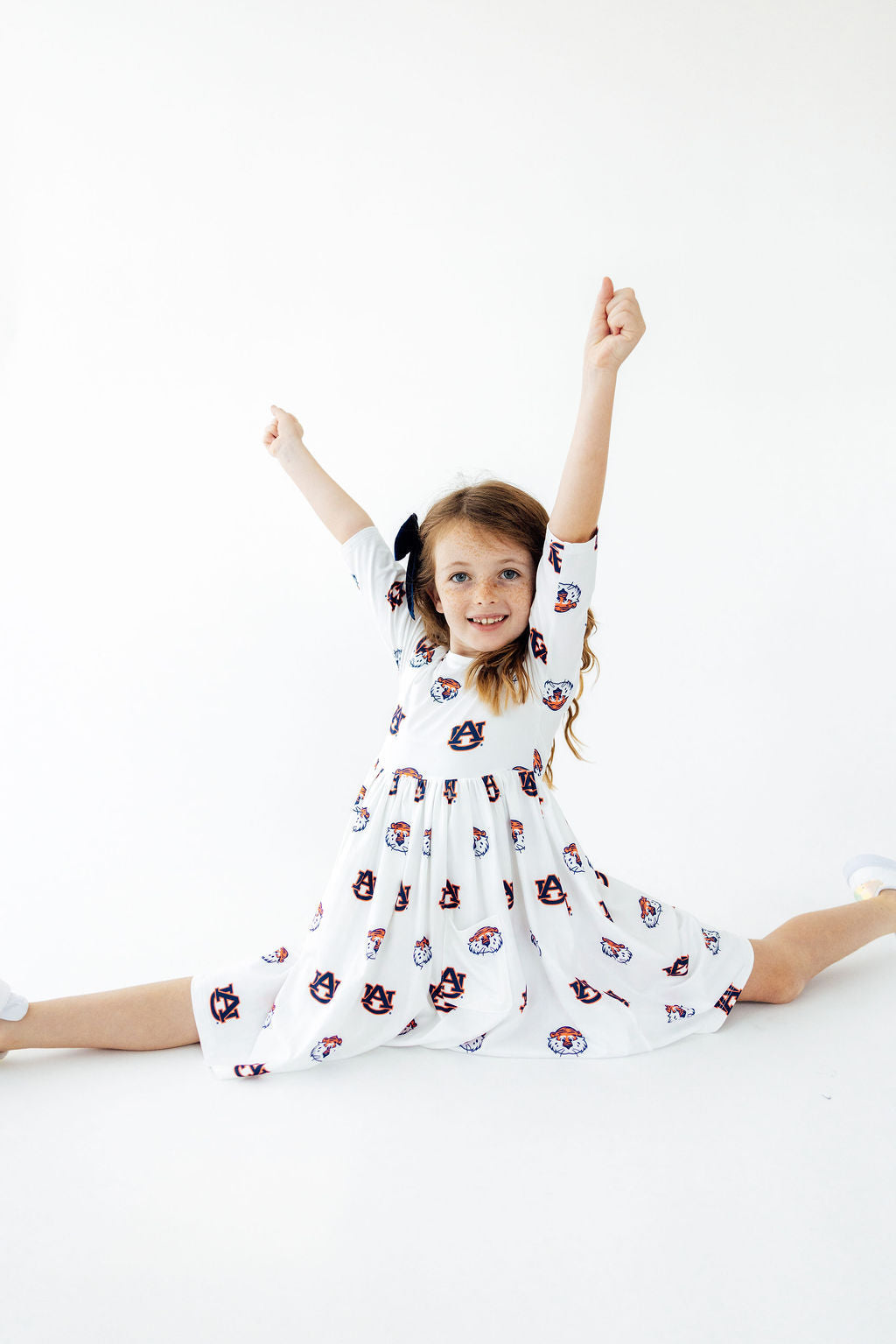 A young girl with brown hair in pigtails sits on the floor in a split position, wearing a white long-sleeve dress with a colorful cartoon cat pattern. She is posing with her arms raised against a seamless white studio background.
