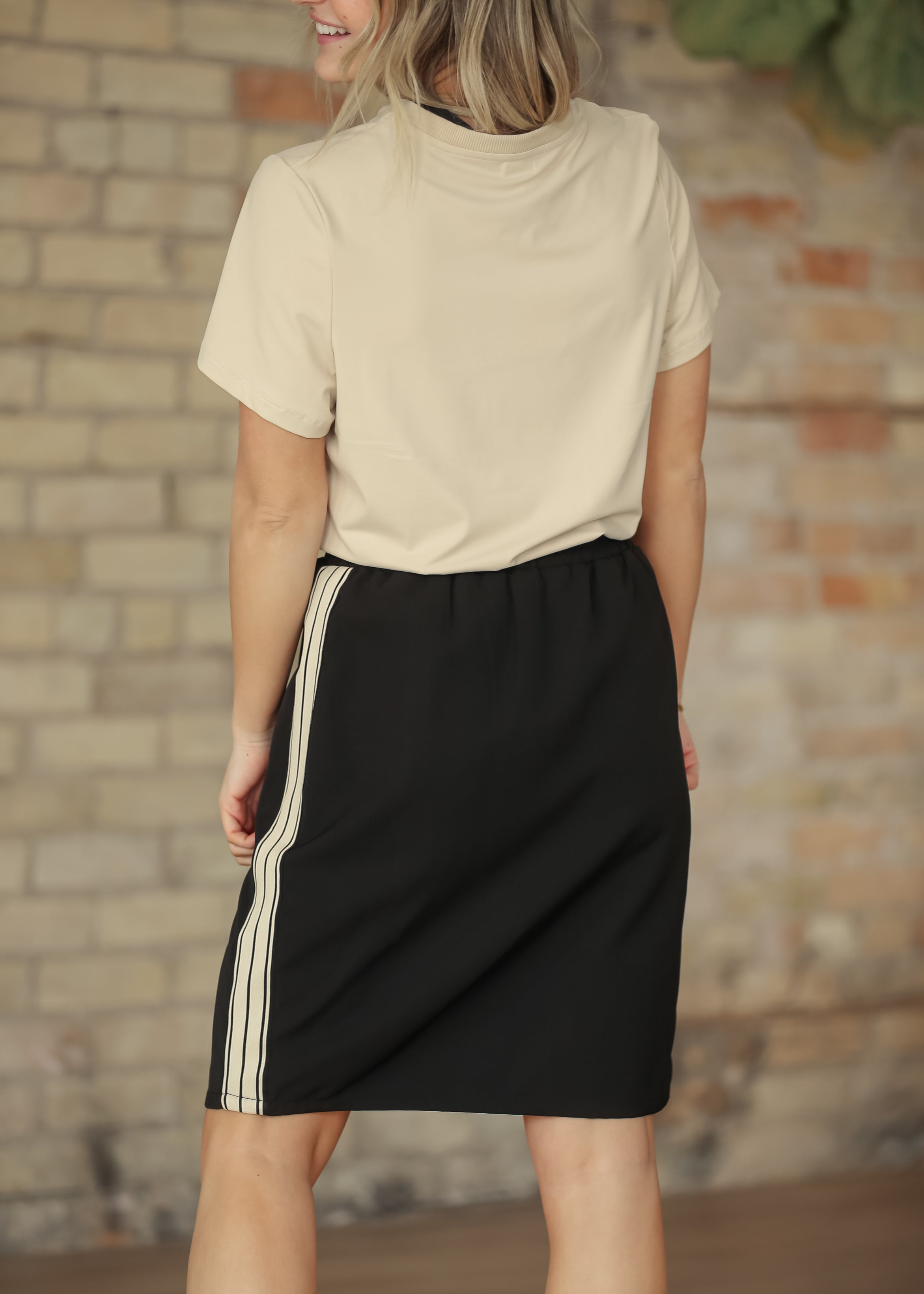 Woman wearing a beige top and black skirt with white stripes against a brick wall.