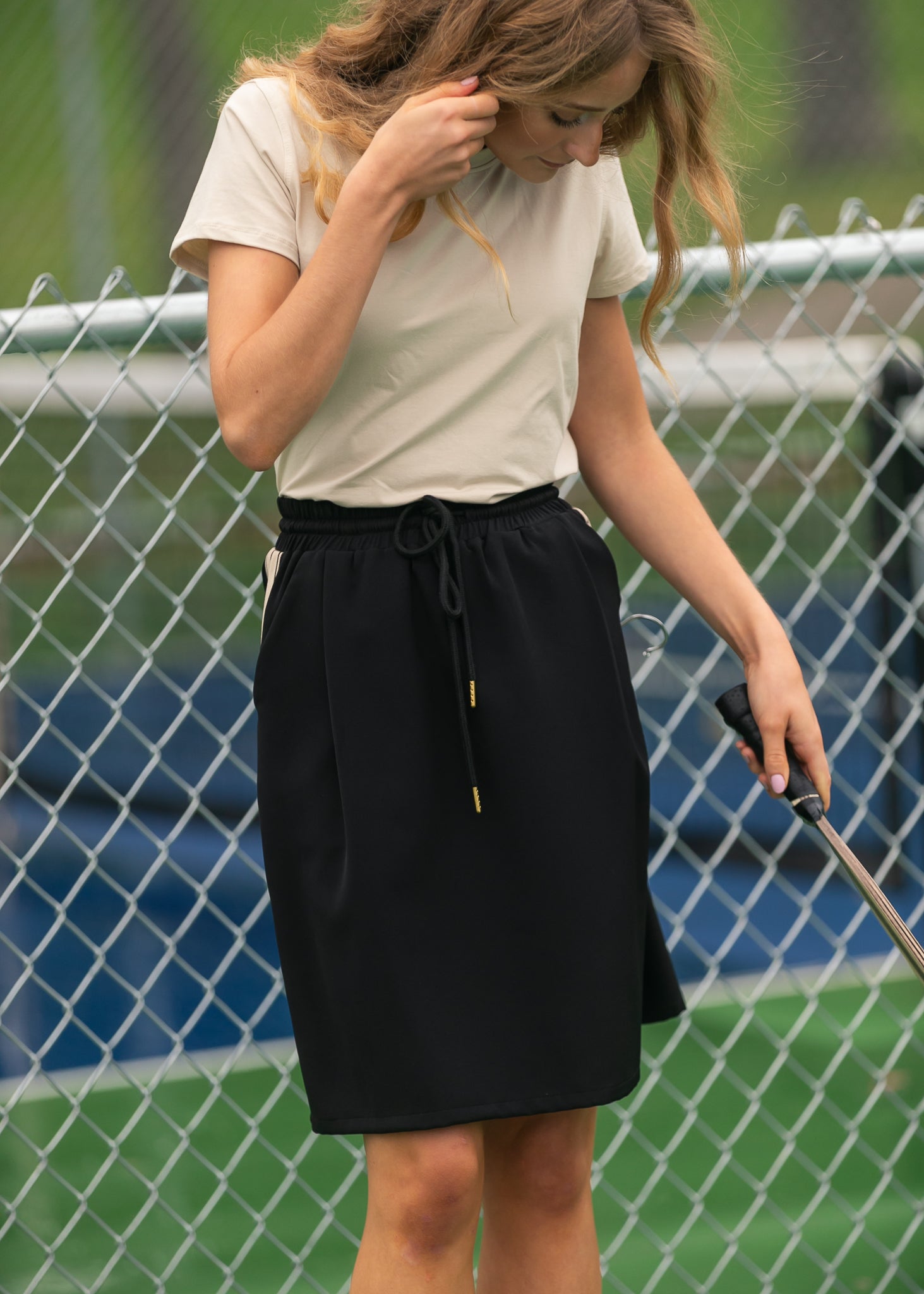 Woman in a white shirt and black skirt holding a golf club, standing in front of a chain-link fence.