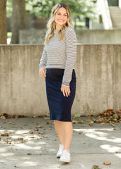 Woman wearing a striped sweater and navy skirt standing outdoors with a concrete wall and trees in the background.
