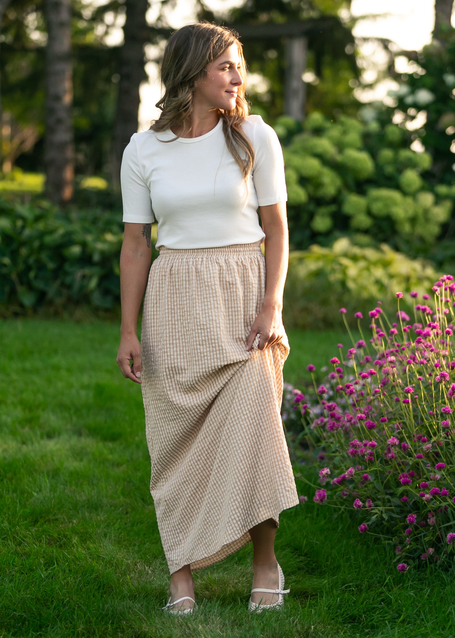 Woman in a white top and beige skirt standing in a garden with greenery and flowers.