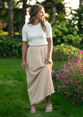 Woman in a white top and beige skirt standing in a garden with greenery and flowers.