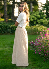 Woman in a white top and beige checkered skirt standing in a garden with greenery and flowers.