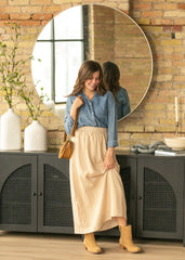 Woman in a blue shirt and beige skirt standing in front of a large round mirror in a room with brick walls and decorative items.