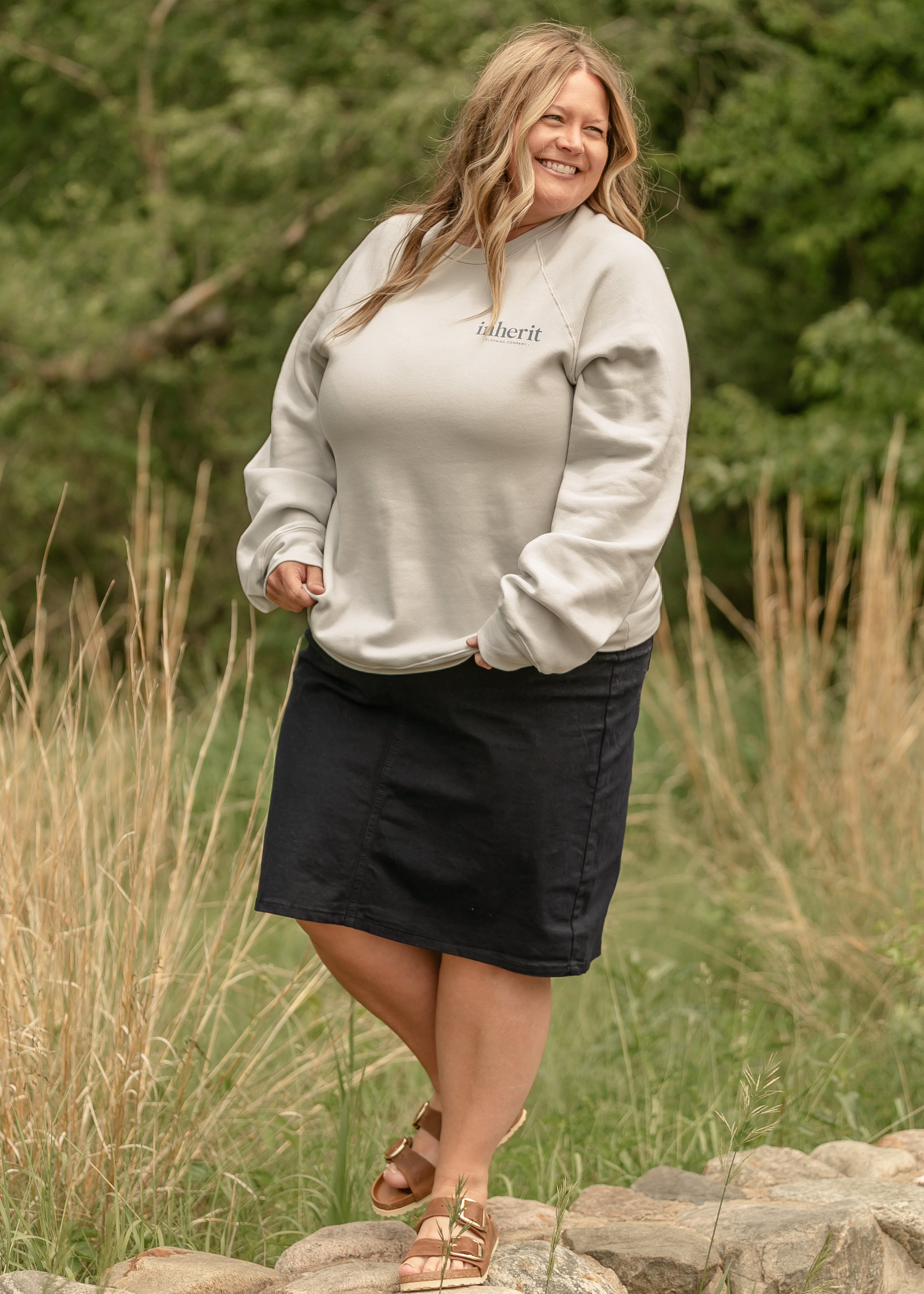 Woman wearing a light gray sweatshirt with a brand logo, standing outdoors in a natural setting.