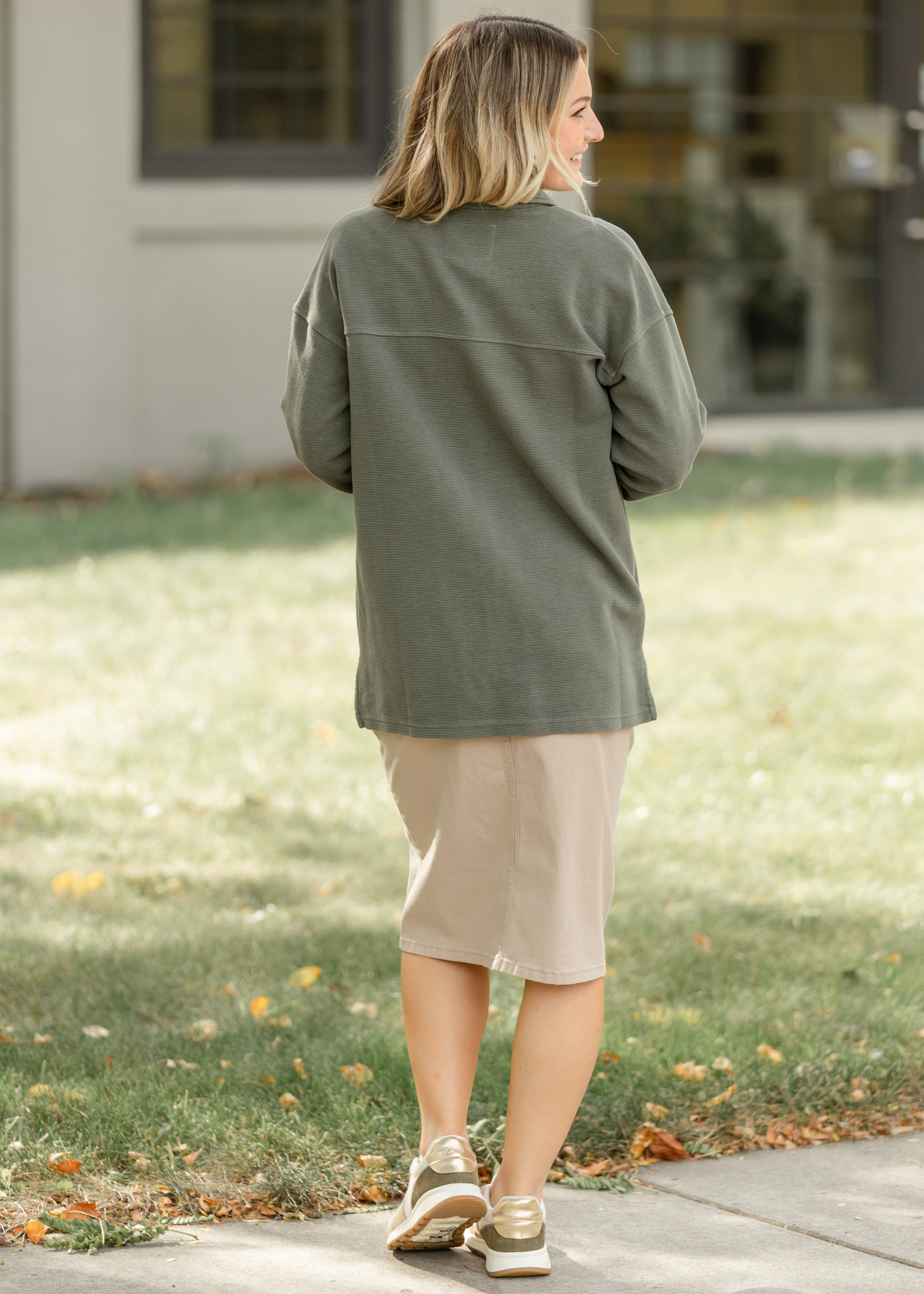 Woman wearing a green jacket and beige skirt walking on a sidewalk.