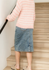 Person wearing a pink striped sweater and blue denim skirt standing on wooden stairs.