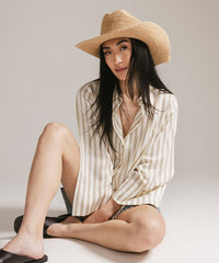 Woman wearing a straw hat and striped shirt sitting on the floor against a plain background