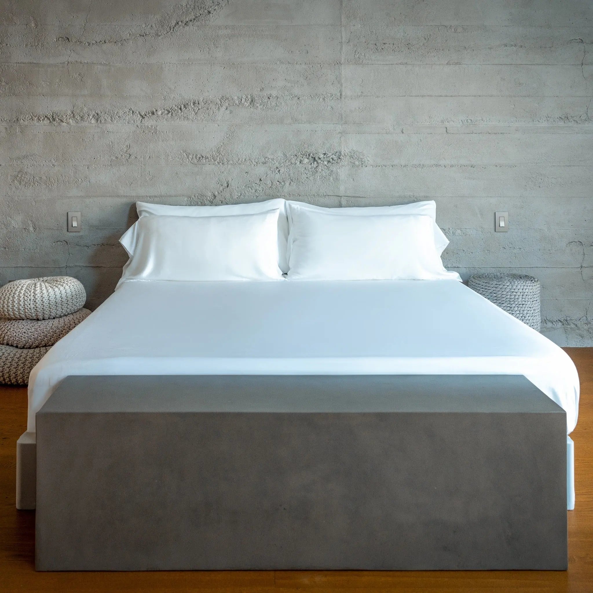 Bed with white bedding and gray mattress cover against a concrete wall.