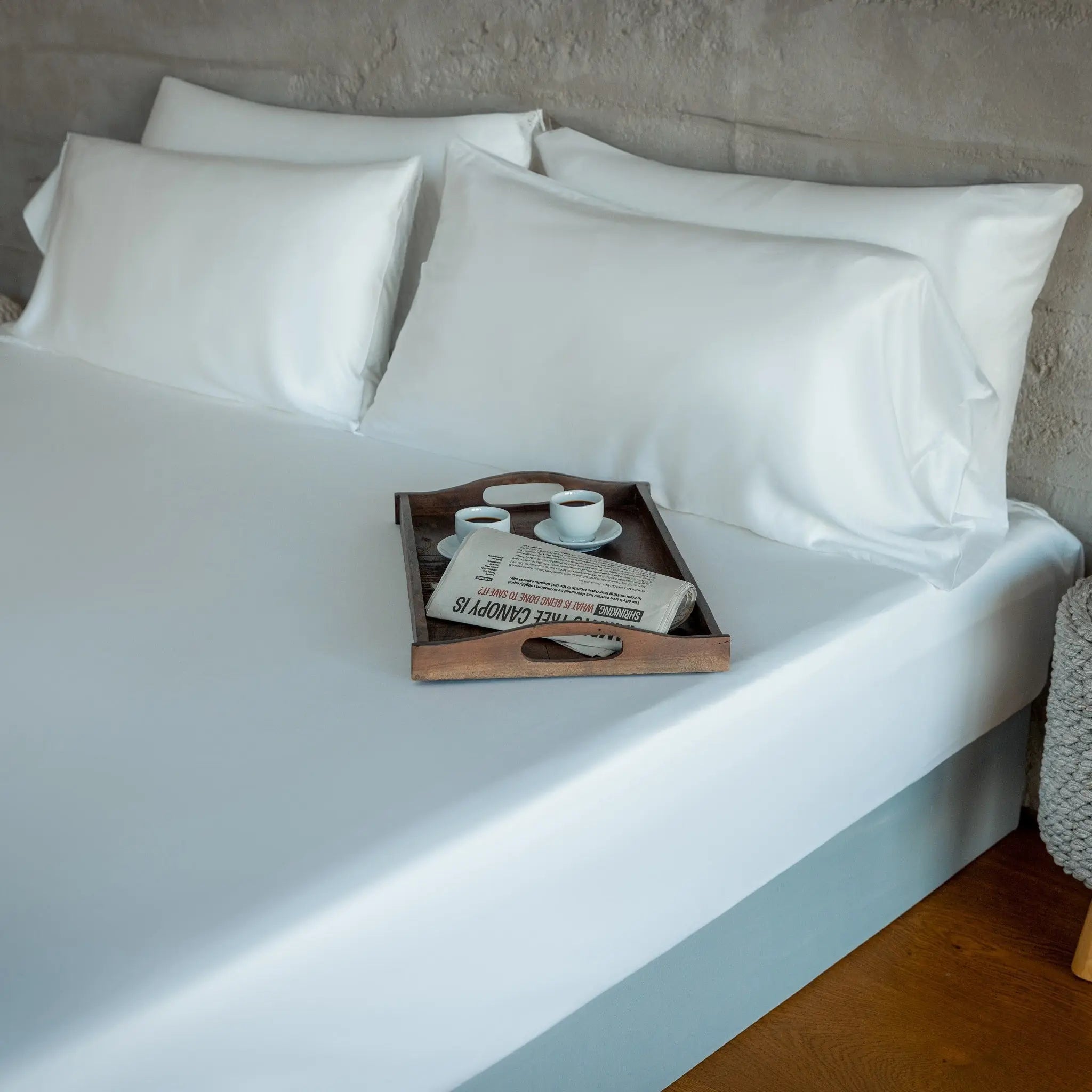 White pillows on a bed with a wooden tray holding cups and a newspaper.
