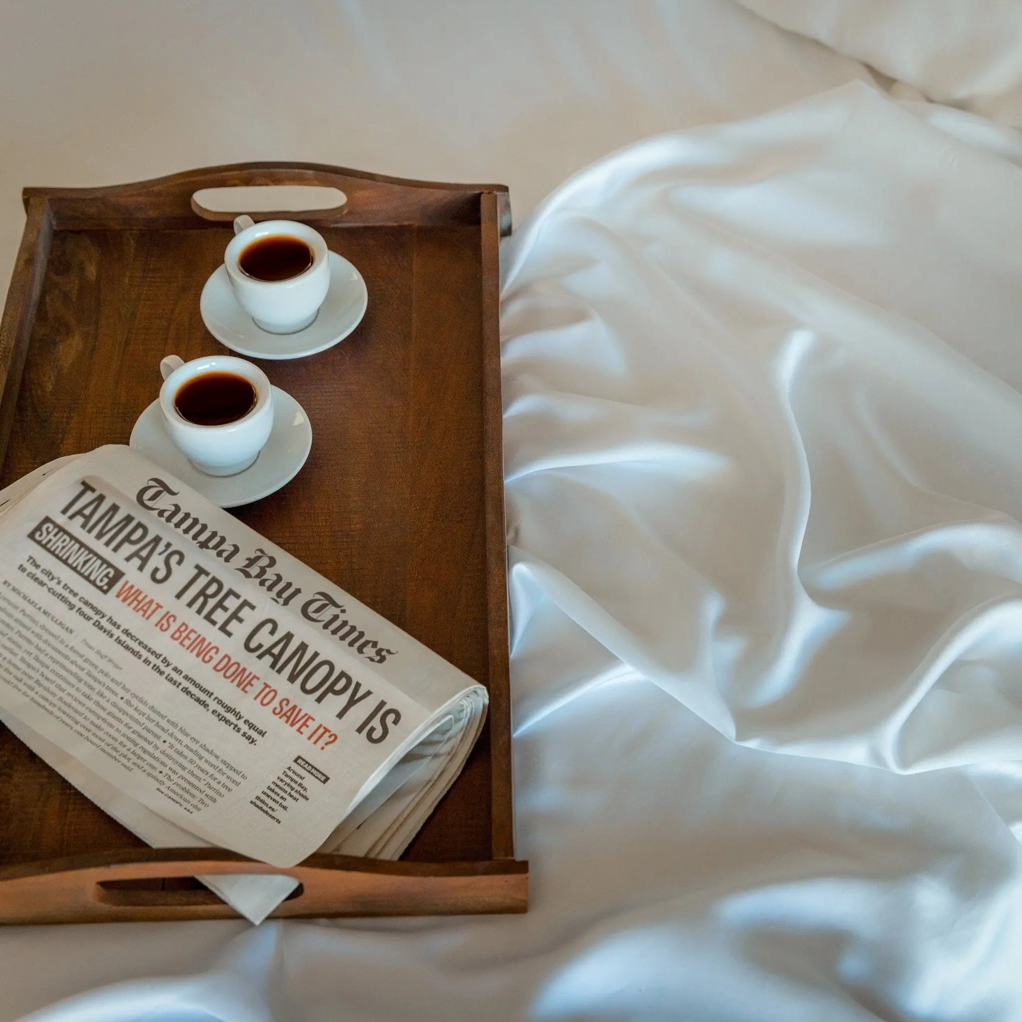 Wooden tray with two cups of coffee and a newspaper on a white bed.