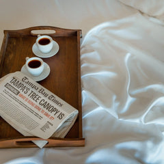 Wooden tray with two cups of coffee and a newspaper on a white bed.