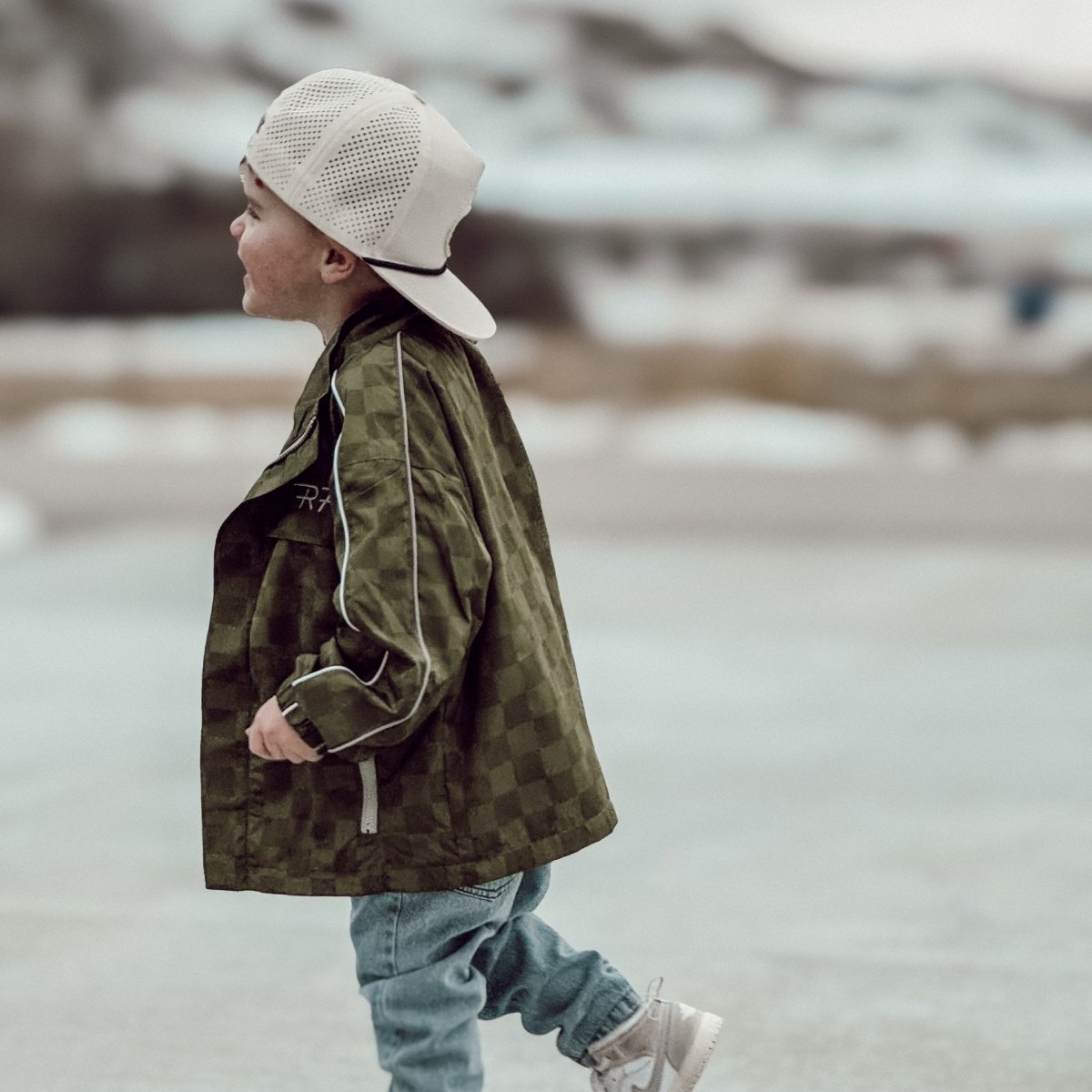 Child wearing a green camouflage jacket and white cap walking on a snowy landscape.