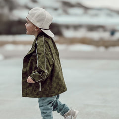 Child wearing a green camouflage jacket and white cap walking on a snowy landscape.