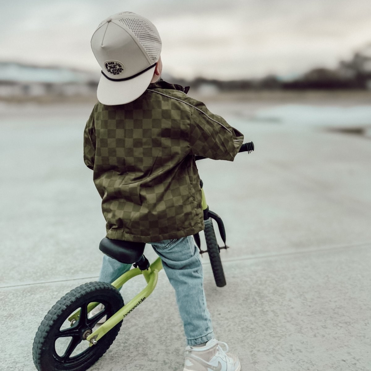 Child in a checkered jacket and cap riding a balance bike on a paved surface.