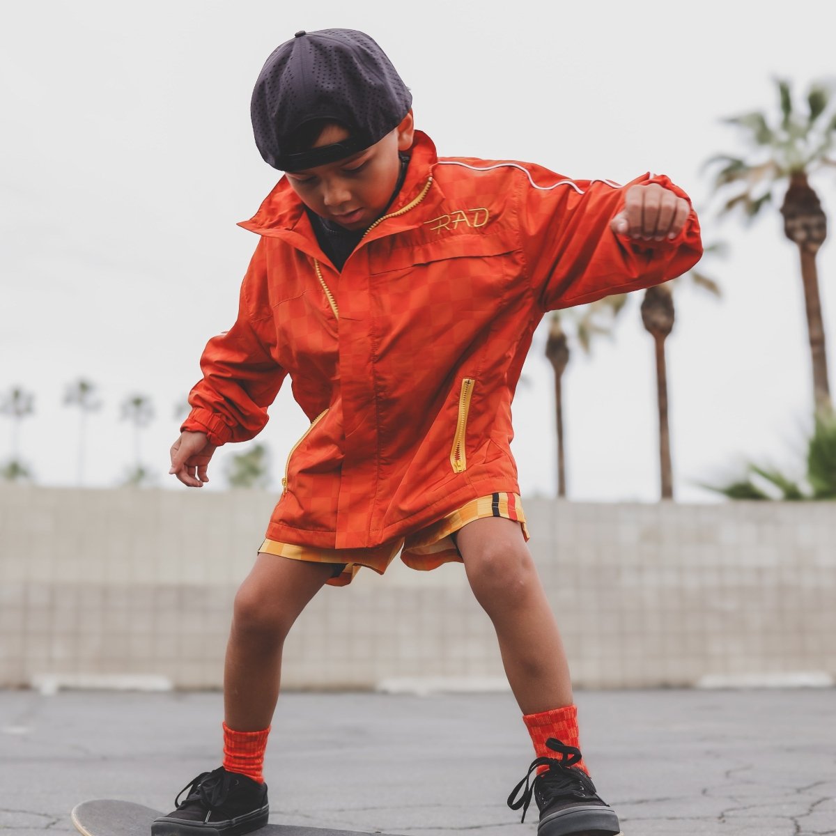 Child in an orange jacket with 'RAD' branding on a skateboard, palm trees in the background