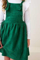Green pinafore dress worn over a white long-sleeve shirt on a plain background