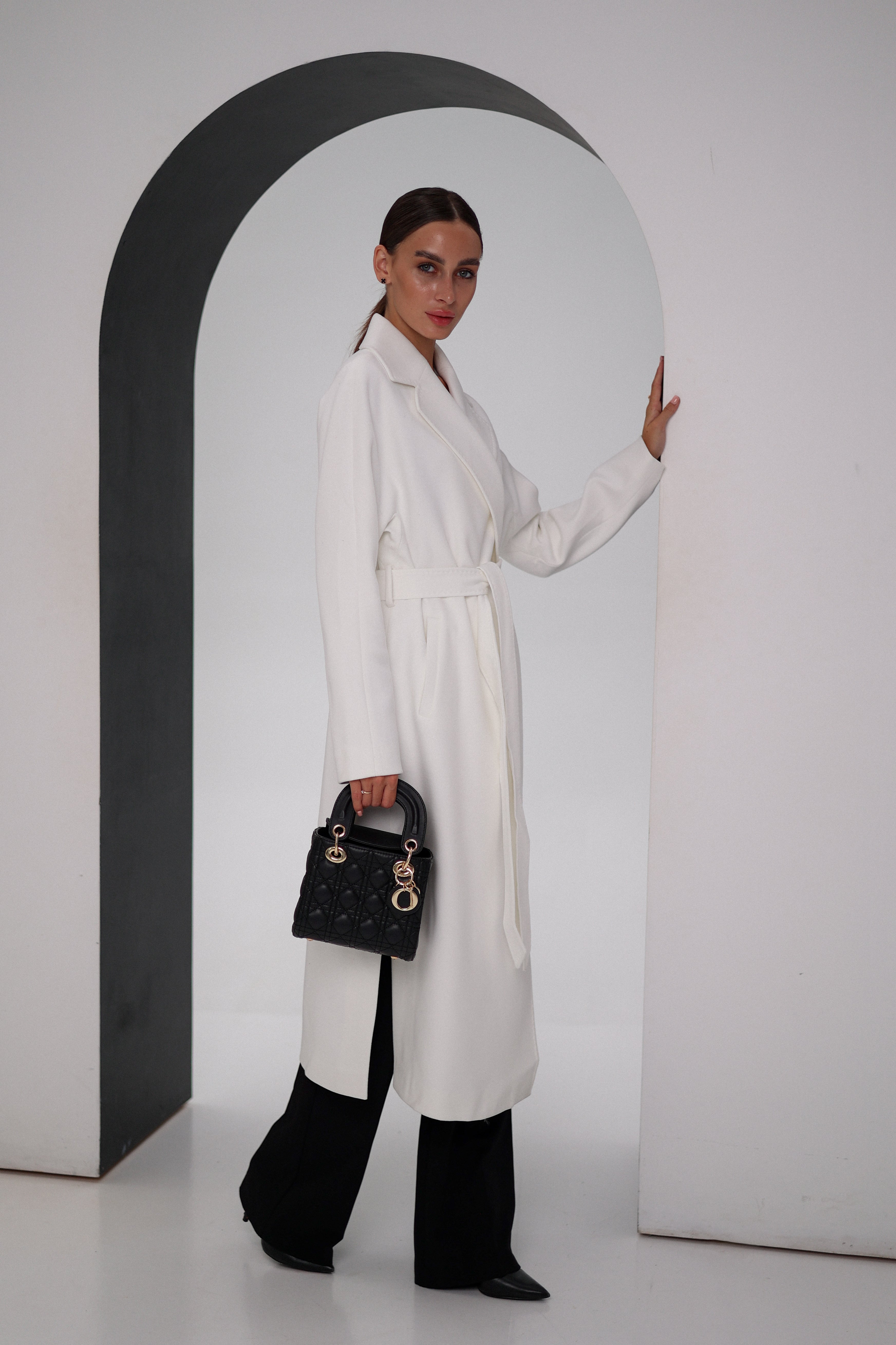 Woman wearing a white coat and black pants, holding a black handbag, against a plain background.