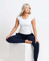 Woman sitting on a white stool wearing a white t-shirt and navy blue pants against a plain background