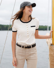 Woman wearing a white shirt with black trim and beige pants by a body of water.