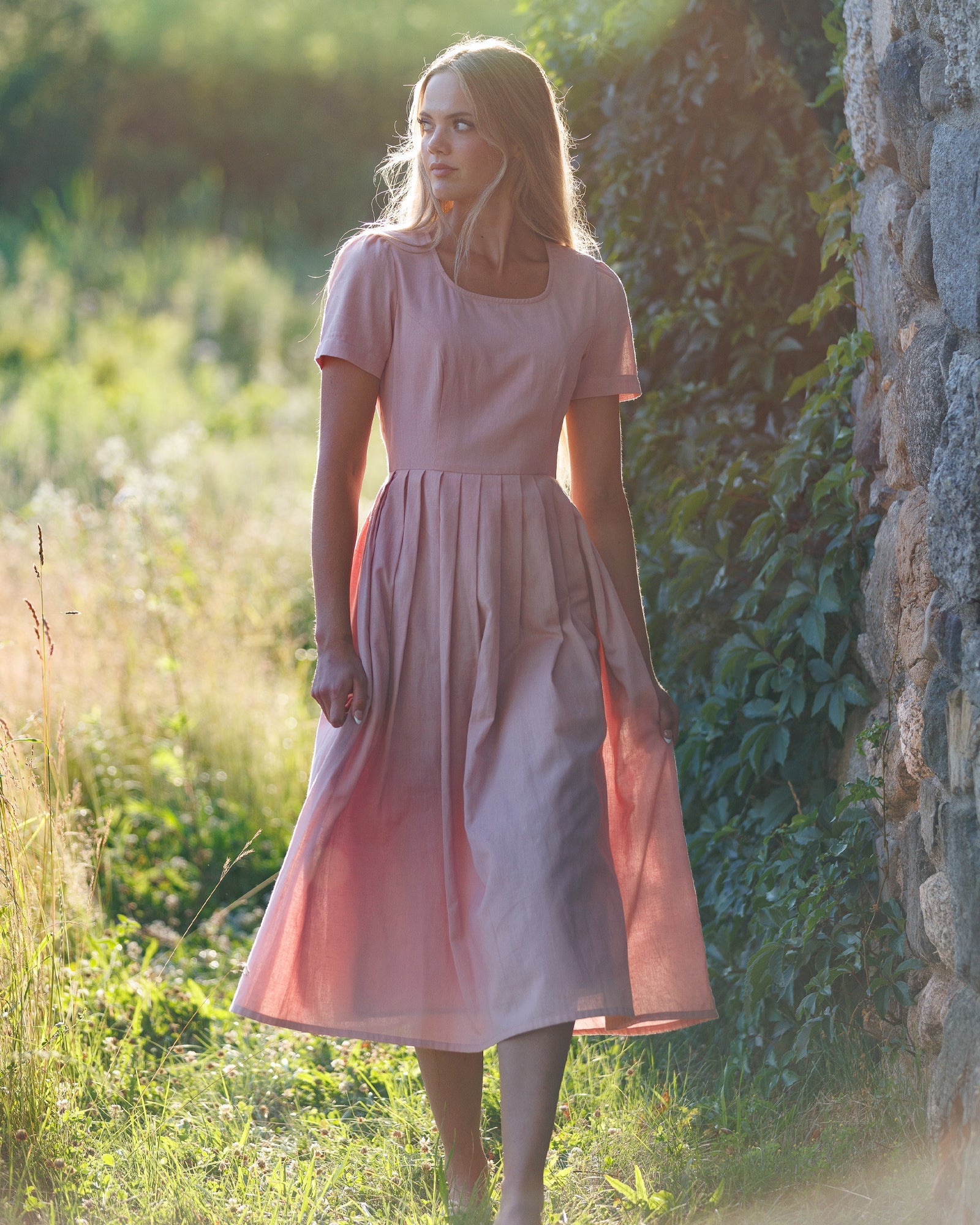 Woman in a pink dress standing in a natural setting with greenery and a stone wall.