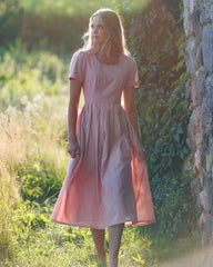 Woman in a pink dress standing in a natural setting with greenery and a stone wall.