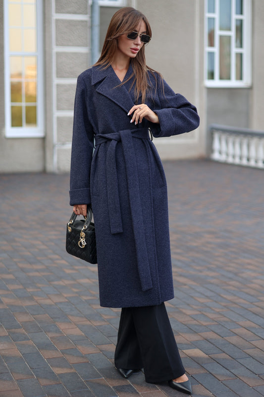 Woman wearing a long navy coat with a belt, holding a black handbag, standing on a paved walkway.