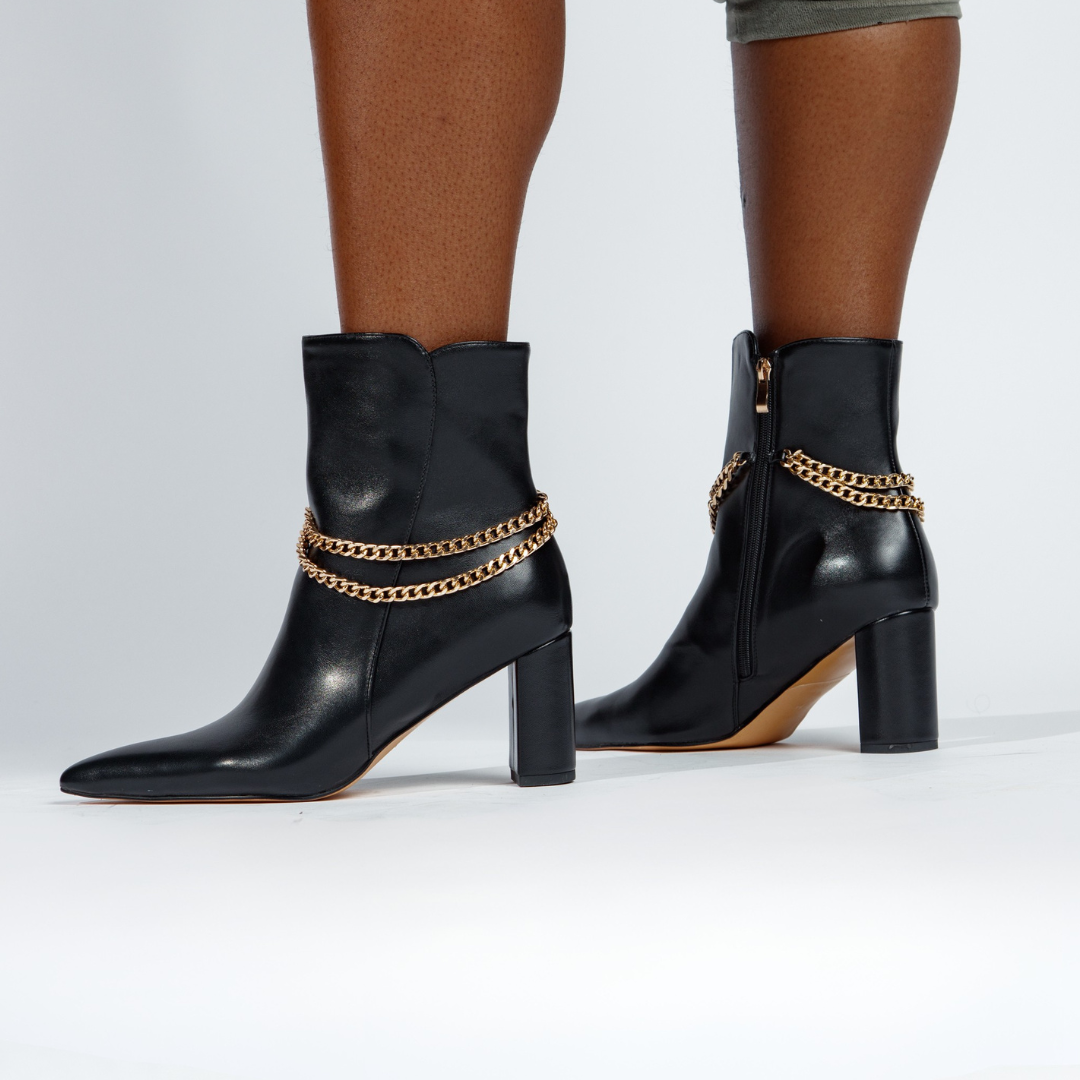 Black ankle boots with gold chain details worn by a person on a white background