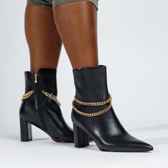 Black ankle boots with gold chain details worn by a person on a white background