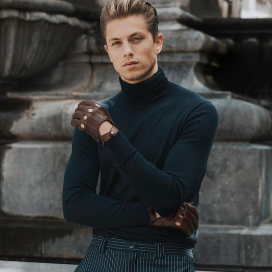 Man wearing a dark turtleneck sweater and brown gloves standing in front of a stone wall.