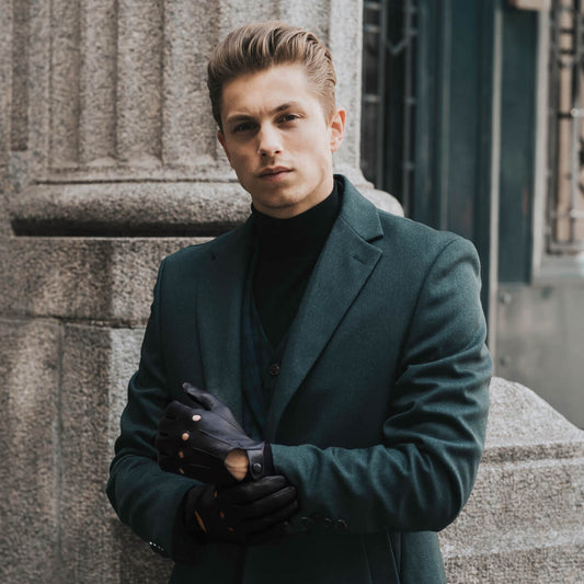 Man in a dark coat and gloves standing against a stone wall.