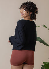 Woman wearing a black long-sleeve top and maroon shorts against a beige background with a plant.